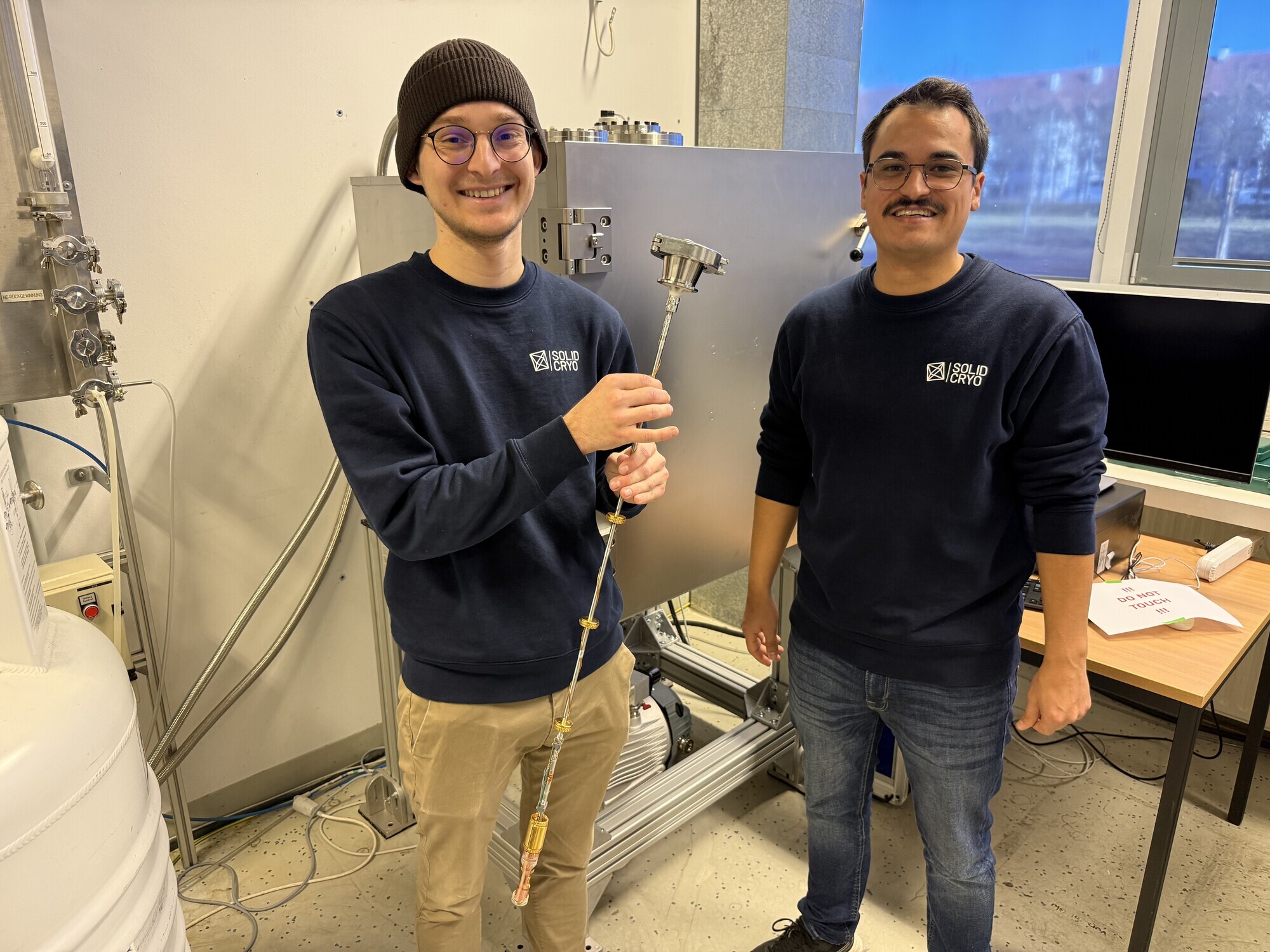 Paul Bittner und Jorginho Villar Guerrero erklären im Interview die Funktionsweise ihrer magnetischen Kühltechnologie. Im Hintergrund steht der Tieftemperatur-Kühlschrank, der für supraleitende Quantencomputer eingesetzt werden soll. Fotoquelle: B4BSCHWABEN.de / Angelina Märkl