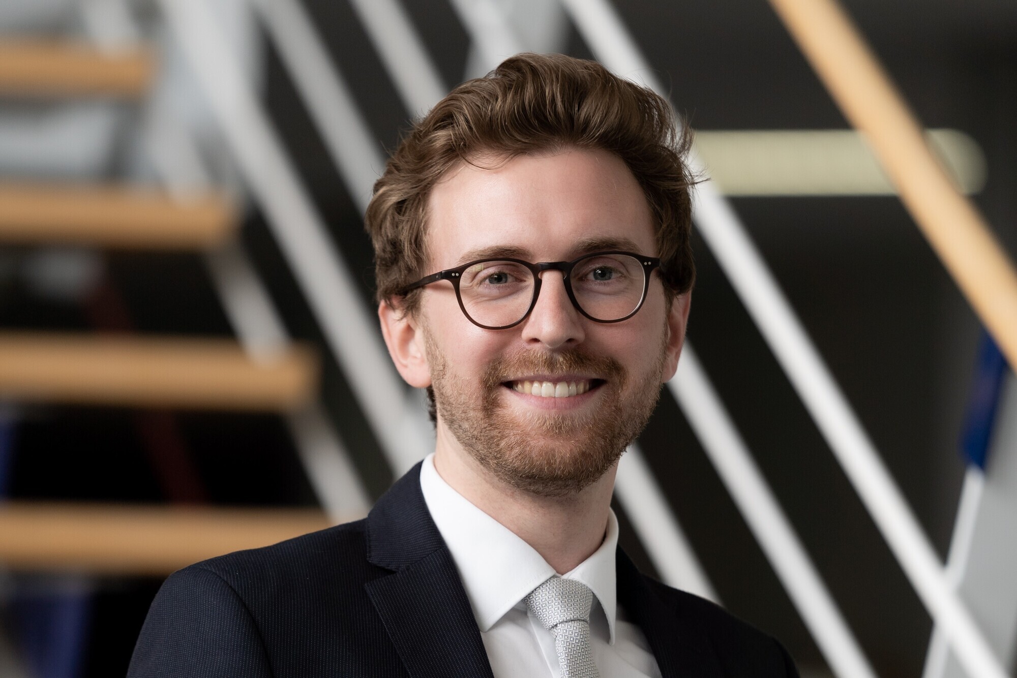 Dr. Moritz Bruckner lächelt in die Kamera. Er hat einen Anzug und eine Brille an.