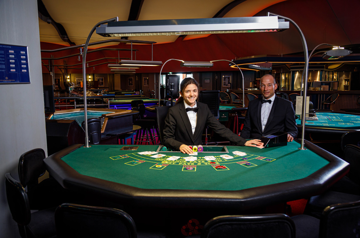 Zwei Croupiers stehen hinter einem Black Jack-Tisch und lächeln in die Kamera