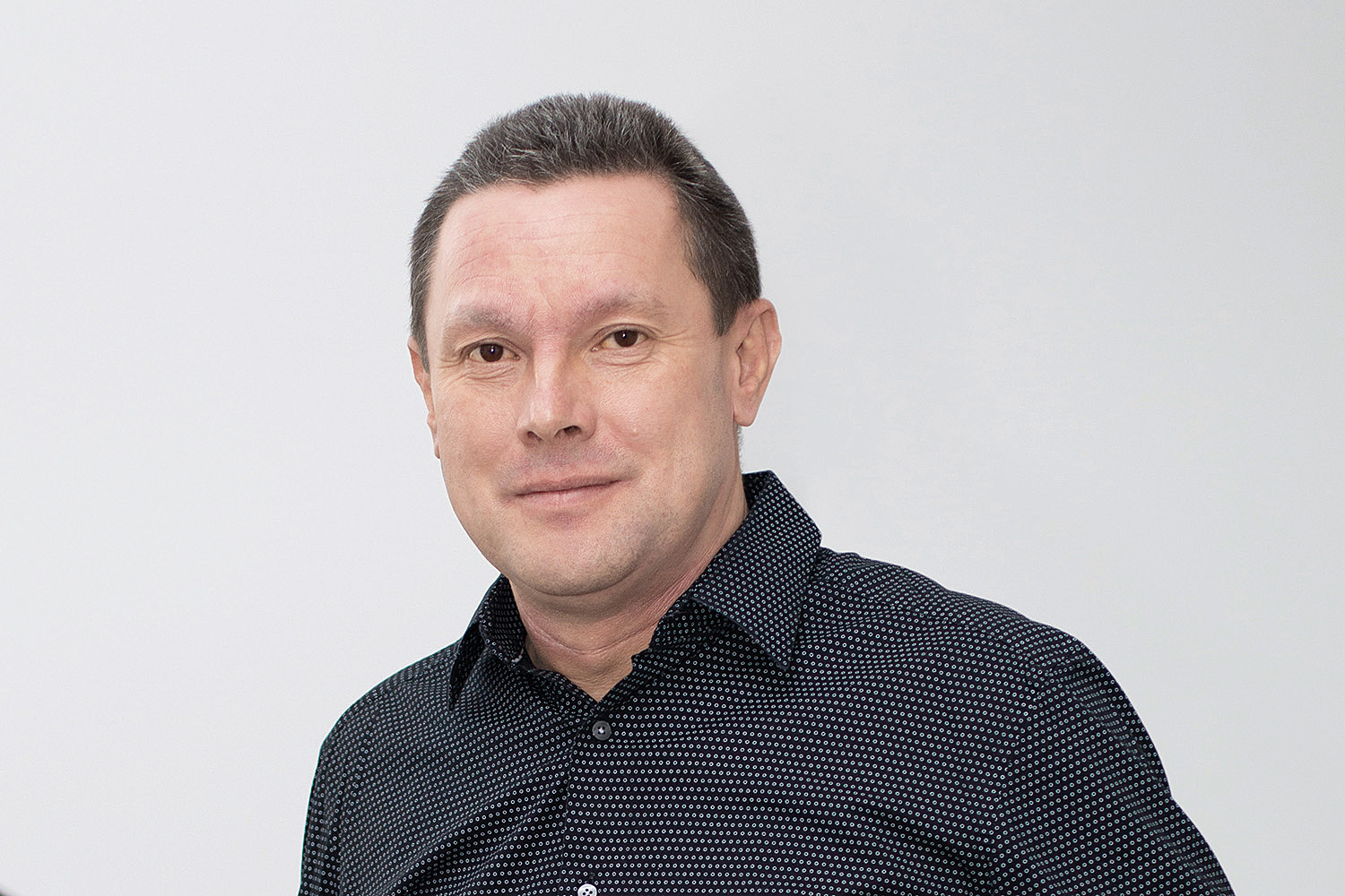 Ein Mann in einem schwarzen Hemd steht vor einer weißen Wand.