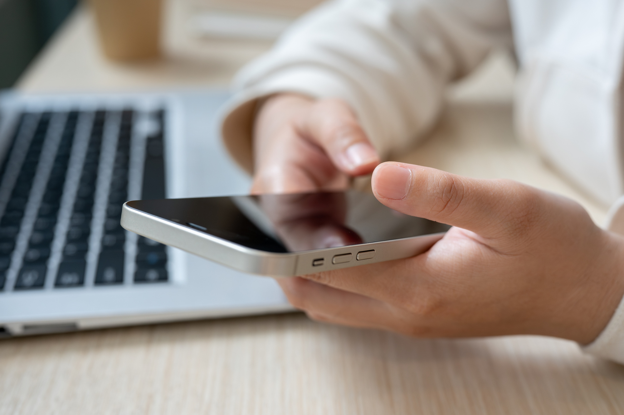 Eine Person hält ein Smartphone in beiden Händen und sitzt vor einem Laptop. Es sind nur die Arme und Hände der Person zu sehen.