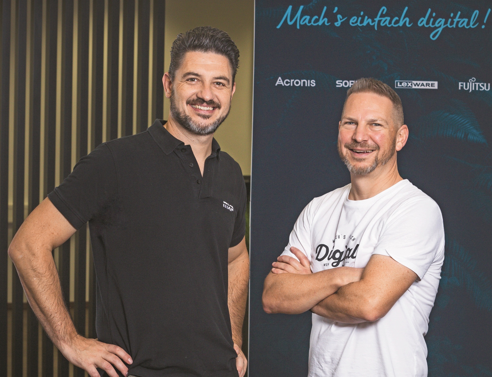 Benjamin Knecht in einem schwarzen T-Shirt steht neben Roland Berger in einem weißen T-Shirt. Beide lächeln in die Kamera