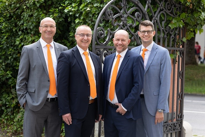 Vier Männer stehen nebeneinander. Sie tragen alle einen Anzug und eine orangene Krawatte. Sie lächeln in die Kamera.