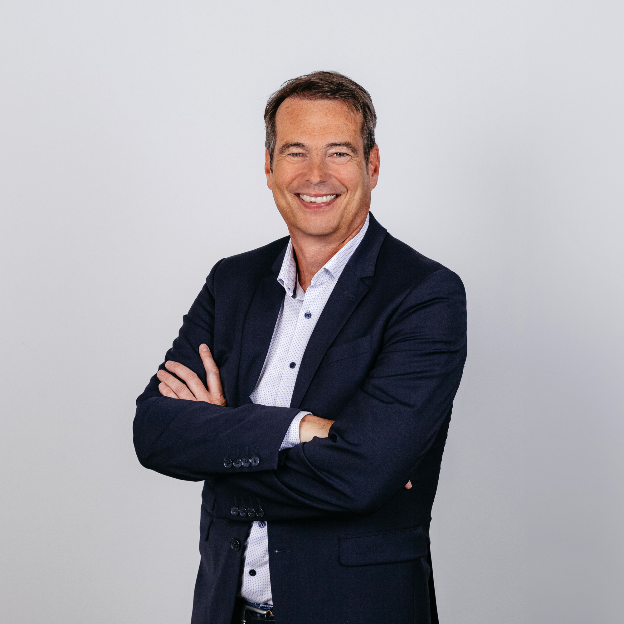 Ein weißer Mann lächelt mit verschränkten Armen in die Kamera. Er trägt einen legeren Anzug ohne Krawatte.